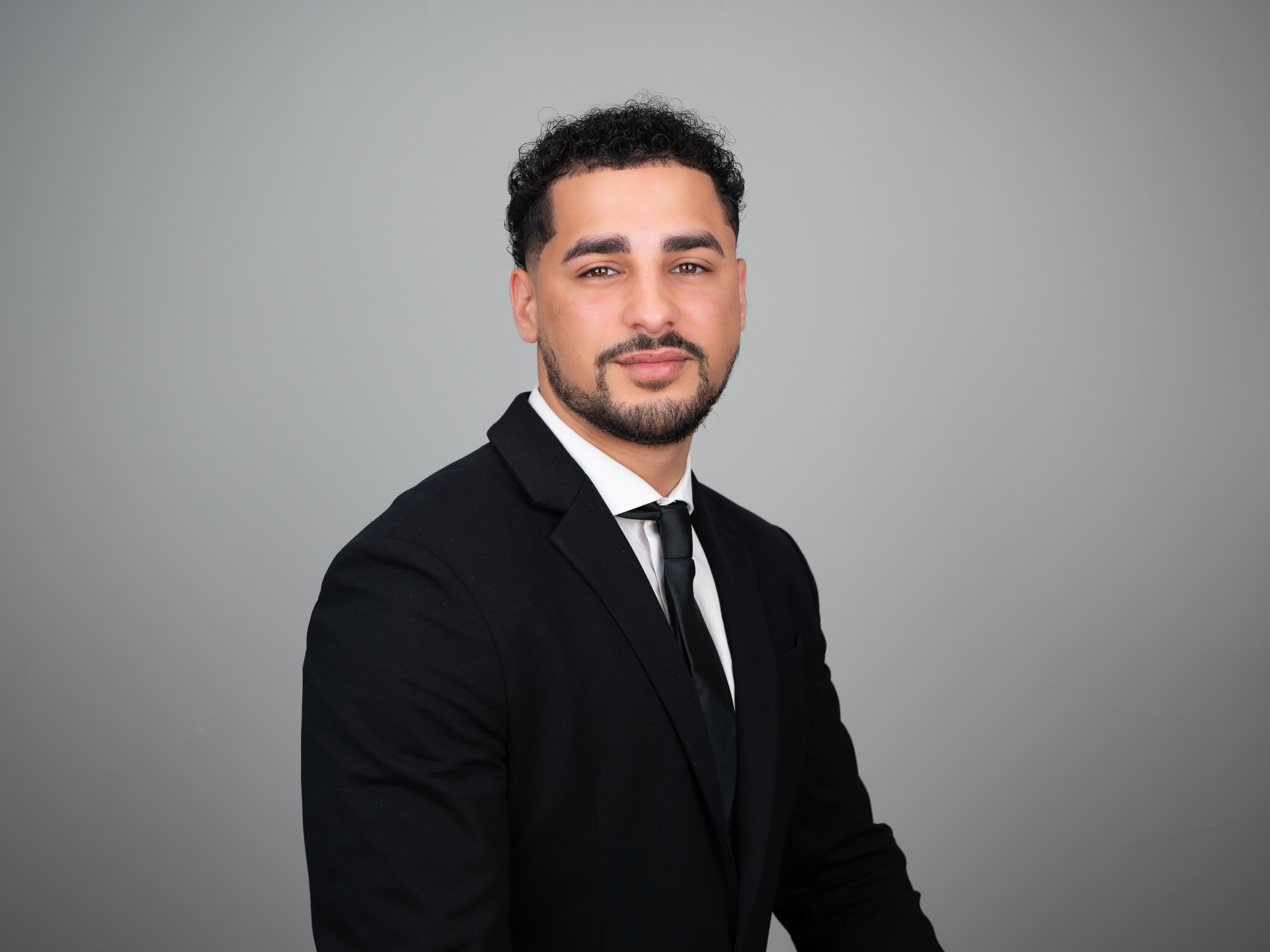 Man with curly dark hair and a beard wearing a black suit and black tie.
